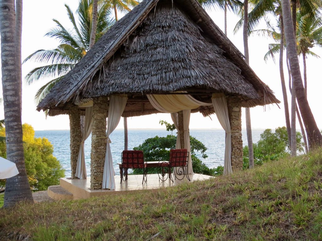 Op vier pilaren rustend puntdak van riet met daaronder twee en een tafeltje met vandaar uitzicht op het strand palmbomen en de indische oceaan op Mafia eiland