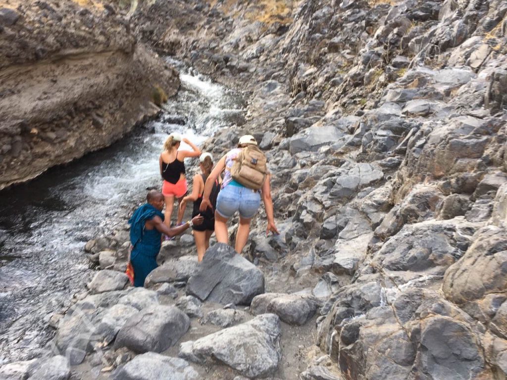 drie vrouwen en een gids op excursie lopend over en tussen de rotsen door naast een riviertje op weg naar een waterval bij Lake Natron