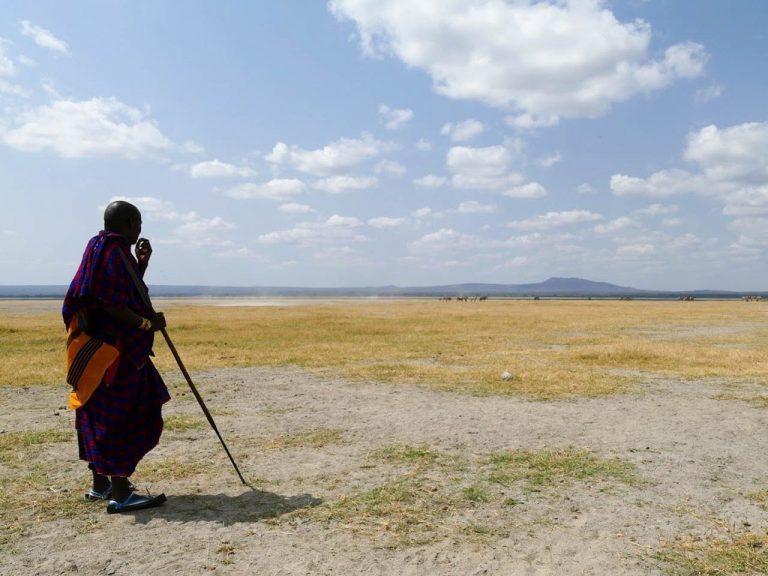 Masai man in traditionele kleding uitkijkend over een droge vlakte met aan de horizon een paar zebra's