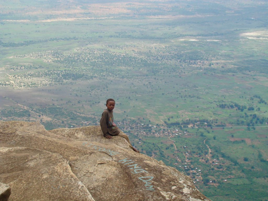 klein jongetje zit op een uitstekende rots heel hoog met uitzicht over de hele groene omgeving beneden hem