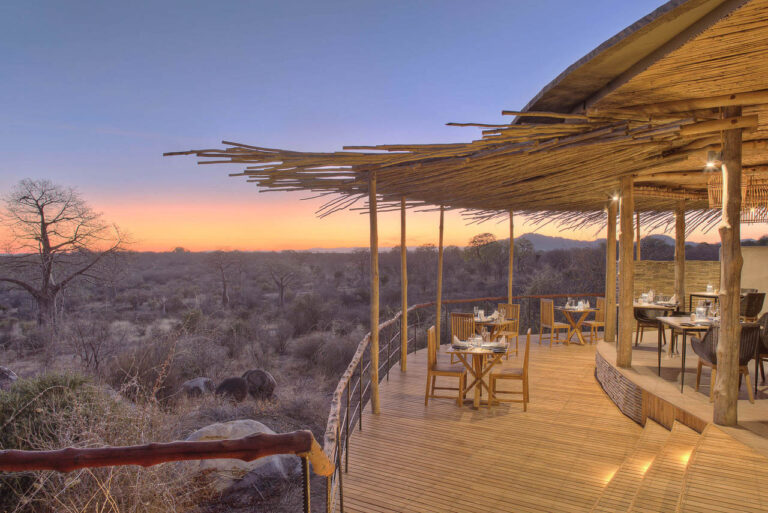 met uitzicht op ruaha een overdekte veranda met tafeltjes aan een open gebouw met meerdere tafeltjes klaar voor het ontbijt in de ochtendschemering
