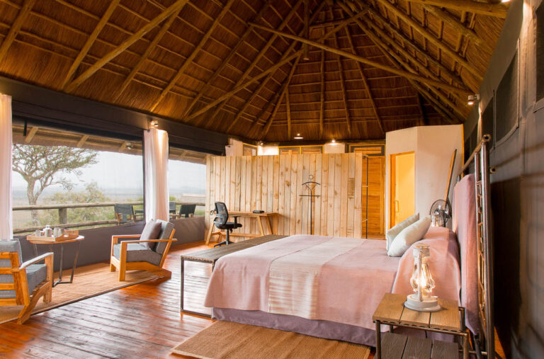 luxe kamer vanuit een zijaanzicht op het mooi opgemaakte bed een hoog puntdak van hout en riet twee comfortabel stoelen en een kleine veranda met uitzicht over tarangire