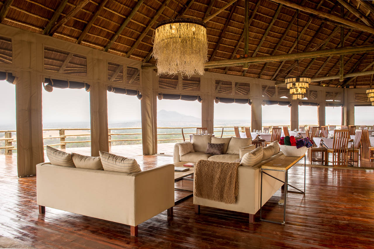 lounge en diner gebouw met op de voorgrond comfortabele witte banken in een u vorm met daarachter gedekte tafels onder een rieten dak met vrij uitzicht over de serengeti