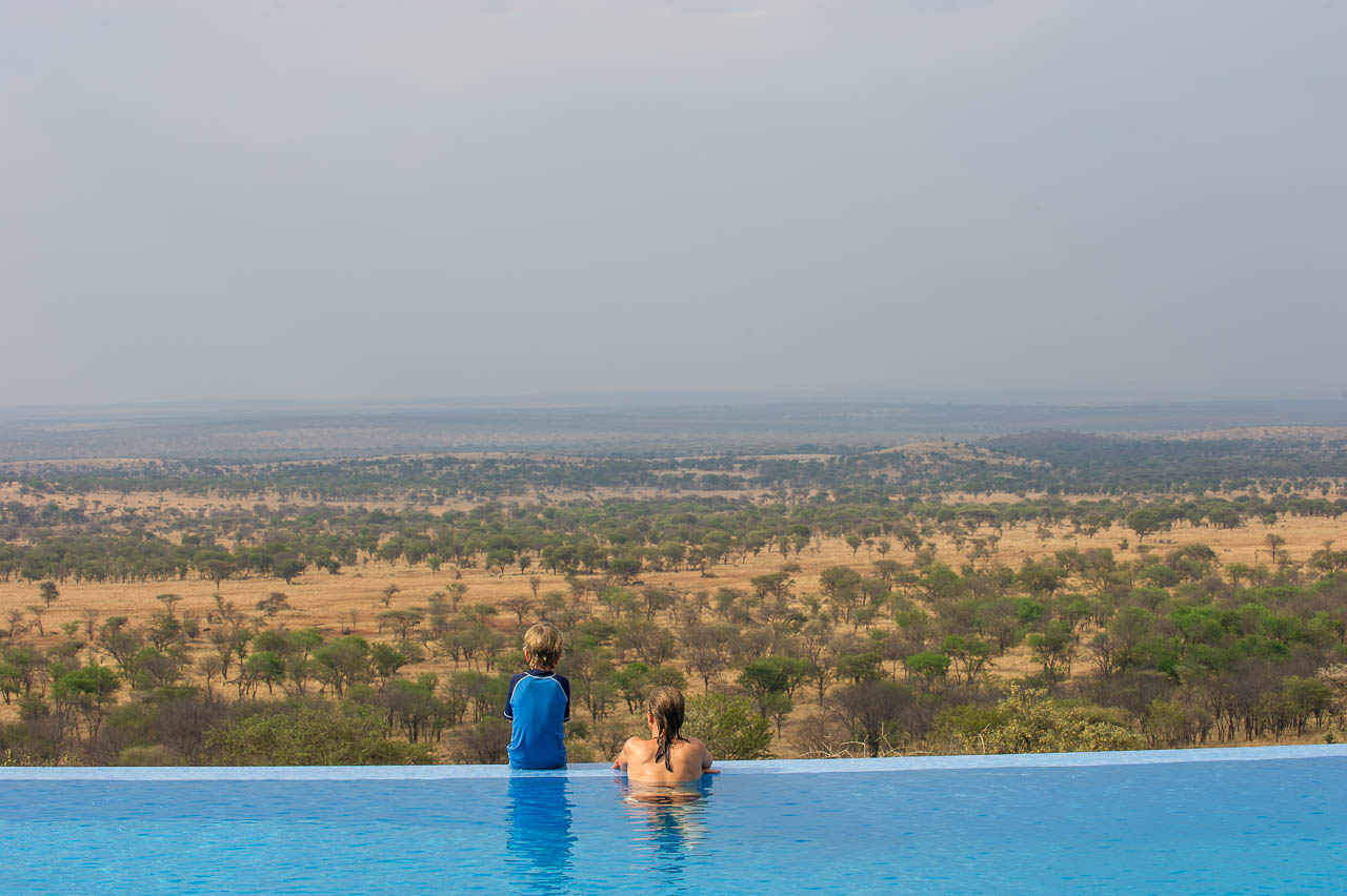 zwembad bij een lodge met jongetje zittend op de rand en vrouw naast hem in het zwembad beide kijkend over de serengeti
