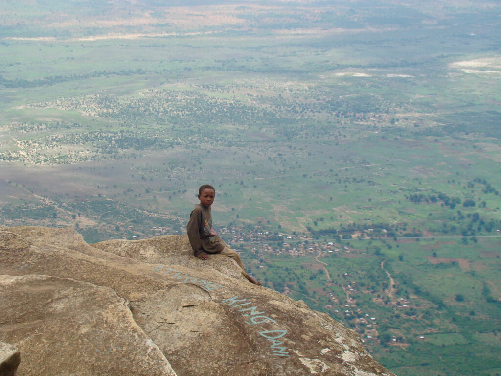 klein jongetje zit op een uitstekende rots heel hoog met uitzicht over de hele groene omgeving beneden hem