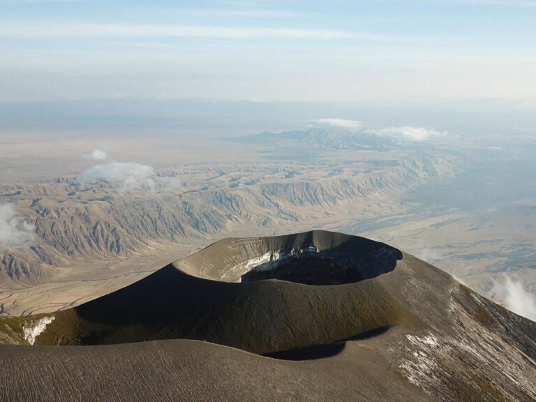 vanuit de lucht de zwarte ronde krater rand van de Ol-doinyo Lengai vulkaan met op de achtergrond een heuvelachtig landschap ontstaan door lava