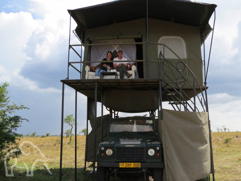 voorkant van een landrovermet recht een douche afgeschermd met canvas een trappetje naar boven waar twee mensen zitten op een bank die staat op een veranda