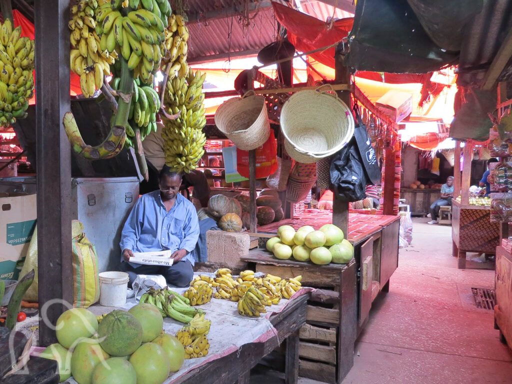 verse meloenen, bananen en manden met zittende lezende man op de Darjani markt Stone Town Zanzibar