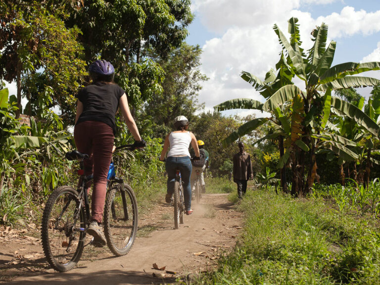 3 mensen fietsend op een mountainbike waarvan een gids op een onverharde weg met bananenbomen recht en een wandelende tanzaniaanse aan de kant