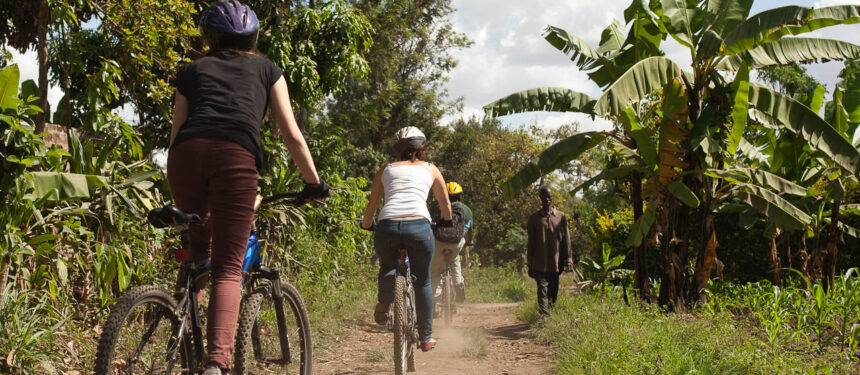 3 mensen fietsend op een mountainbike waarvan een gids op een onverharde weg met bananenbomen recht en een wandelende tanzaniaanse aan de kant