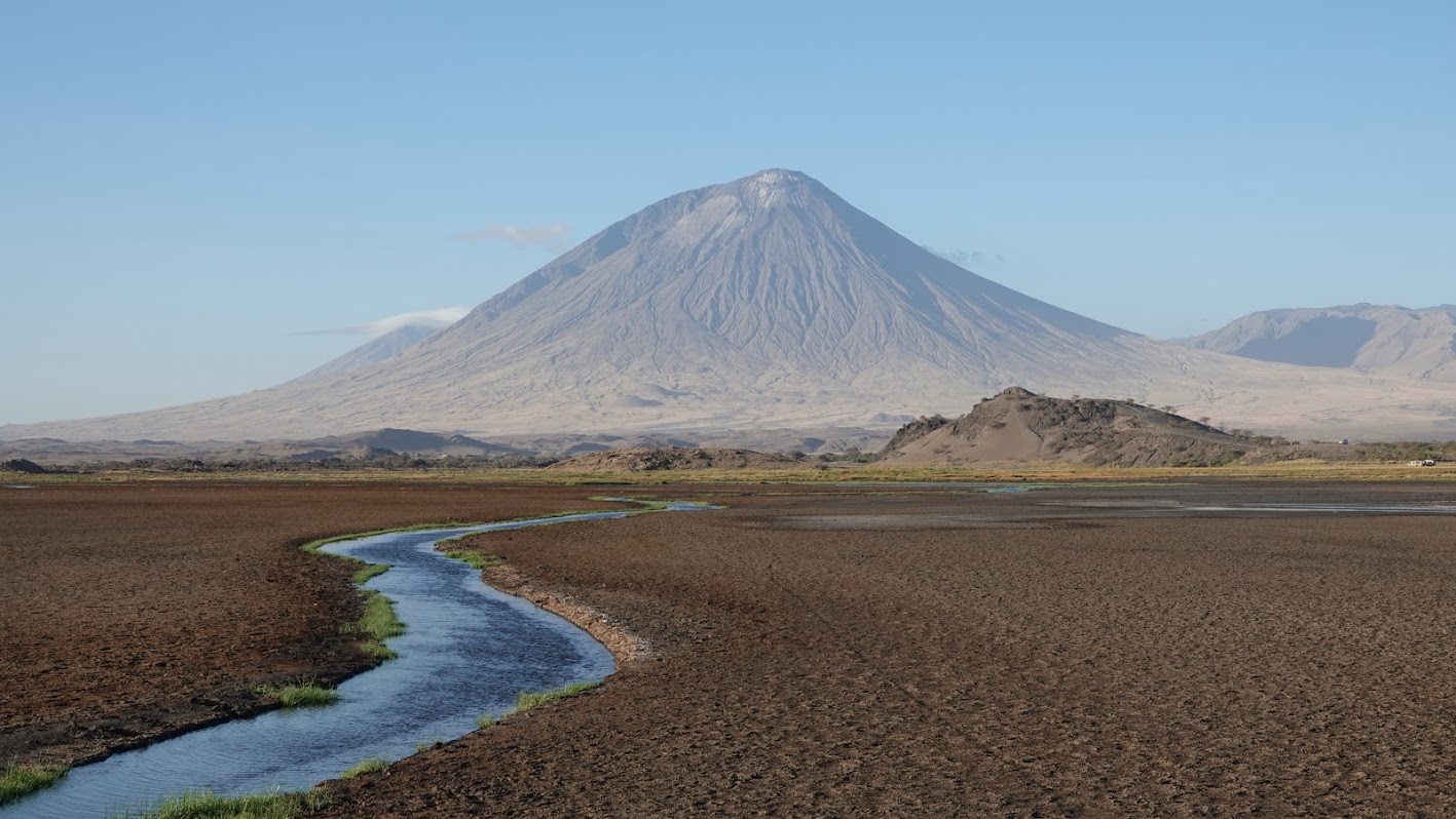 zwarte vlakte met riviertje er door heen met uitzicht op de vulkaan ol doinyo lengai de heilige berg van de masai bij lake natron tanzania