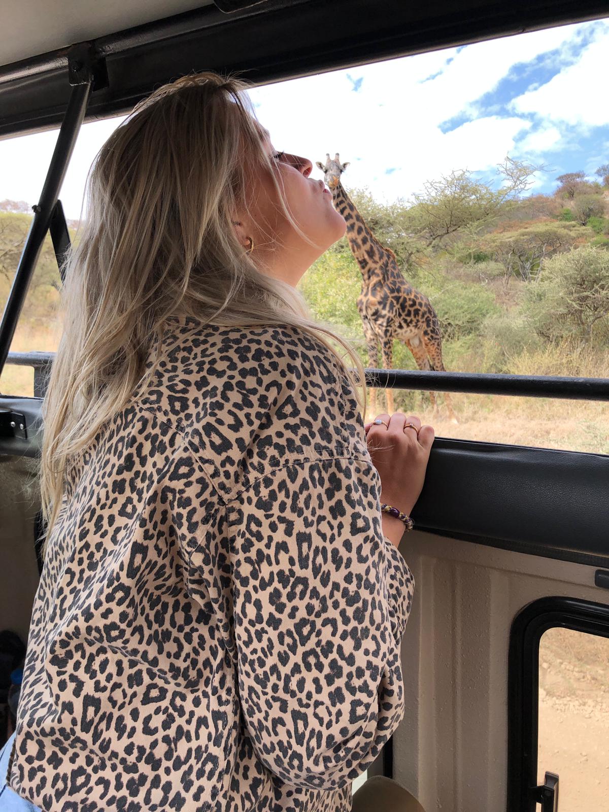 Safari rondreizen door Tanzania ter inspiratie. Er staat een vrouw in de safariauto en op de achtergrond staat een giraf. De foto is zo gemaakt dat het lijkt of de vrouw de giraf kust.