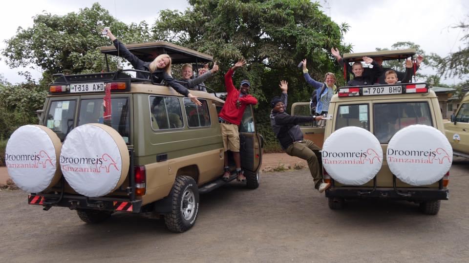 Reiservaringen rondreis Tanzania. Gasten van droomreis Tanzania met hun gidsen die allemaal blij kijken en ze hebben de handen omhoog. Hangen deels aan en uit de auto. Ook staan ze in de auto onder het opendak.