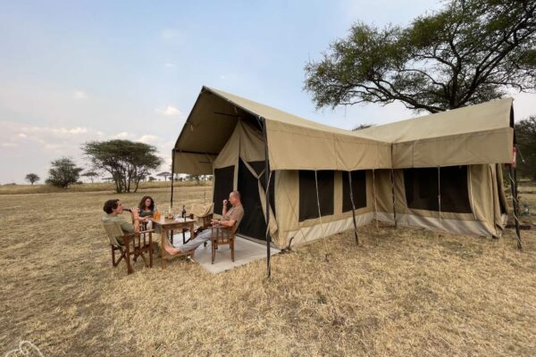 Serengeti rondreizen in Tanzania ter inspiratie. Op de vlaktes van de Serengeti staat een safari tent waarvoor 3 mensen zitten te borrelen.