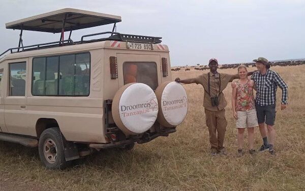 op de voorgrond de safari-auto van droomreis tanzania met daarnaast de gids en twee gasten tijdens hun huwelijksreis. op de achtergrond tot aan de horizon een grote groep gnoes tijdens de grote migratie op de serengeti