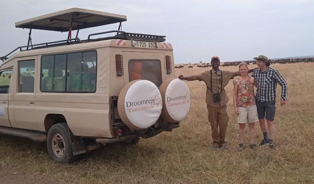 op de voorgrond de safari-auto van droomreis tanzania met daarnaast de gids en twee gasten tijdens hun huwelijksreis. op de achtergrond tot aan de horizon een grote groep gnoes tijdens de grote migratie op de serengeti