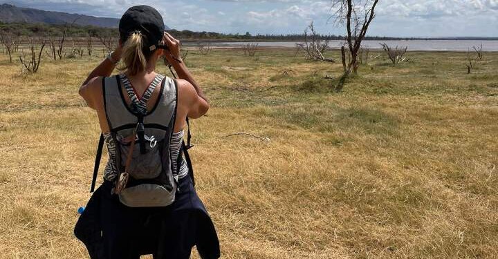 Safari tanzania. Tijdens een wandelsafari kijkt een vrouw met de verrekijker over de goudgele vlaktes met daarachter een meer of ze iets kan spotten