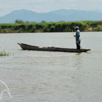 visser visser staand op een klein smal houten bootje op een rivier met aan de oevers veel groen en verderop een berglandschap bij Udzungwa