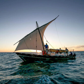 dhow dhow met mast en man daarnaast met geheven zeil en overdekt zitgedeelte met een stuurman daarachter varend op de indische oceaan bij Fanjove