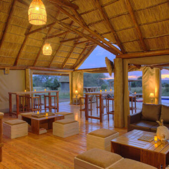 in een luxe lodgebij de loungeruimte twee comfortabele banken met links daarachter twee bartafels en een zwembad bij een ondergaande zon