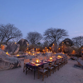 in ruaha bij schemering tussen allemaal rotsblokken met lampjes erop een grote gedekte tafel met kaarsjes klaar voor het diner