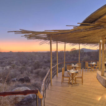 met uitzicht op ruaha een overdekte veranda met tafeltjes aan een open gebouw met meerdere tafeltjes klaar voor het ontbijt in de ochtendschemering