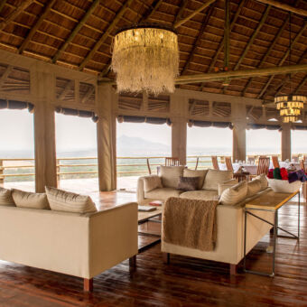 lounge en diner gebouw met op de voorgrond comfortabele witte banken in een u vorm met daarachter gedekte tafels onder een rieten dak met vrij uitzicht over de serengeti