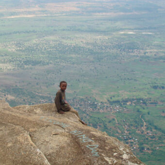 vergezicht klein jongetje zit op een uitstekende rots heel hoog met uitzicht over de hele groene omgeving beneden hem