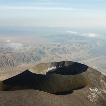 de heilige berg van de Masai vanuit de lucht de zwarte ronde krater rand van de Ol-doinyo Lengai vulkaan met op de achtergrond een heuvelachtig landschap ontstaan door lava