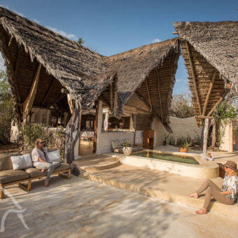 de mooiste locaties twee mensen zittend waarvan 1 op een bank en de ander tegen paal op de grond bij een bar en vijver behorend bij een luxe accomodatie in het zuider van tanzania