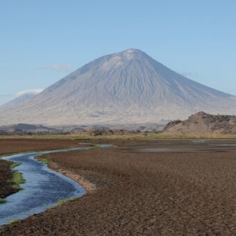 zwarte vlakte met riviertje er door heen met uitzicht op de vulkaan ol doinyo lengai de heilige berg van de masai bij lake natron tanzania