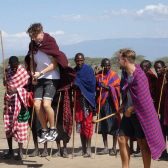 kleurrijke masai mannen die in een kringetje staan en waarvan twee springen en een wedstrijdje doen met gasten van droomreis tanzania om te kijknen ie het hoogst kan springen