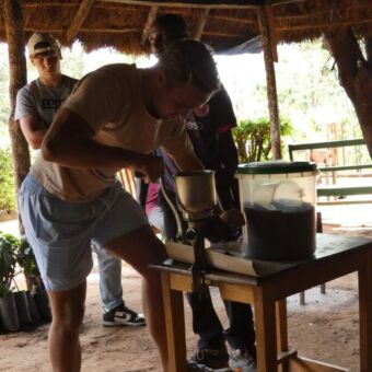 2 gasten van droomreistanzania die proberen koffie te malen. de gids kijkt toe hoe het gaat.