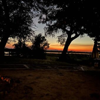 zonsondergang met op de voorgrond een kampvuur en daarachter bossages en dan een oranje horizon