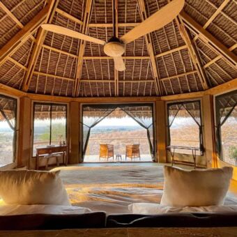 In een grote luxe kamer met veel door zichtige gazen wanden een groot bed onder een hoog schuin rieten dak en ervoor een veranda met 2 stoelen