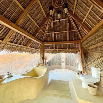 Luxe badkamer zonder wanden met een schuin rieten dak en met een bad, wastafel, toilet en een buitendouche.