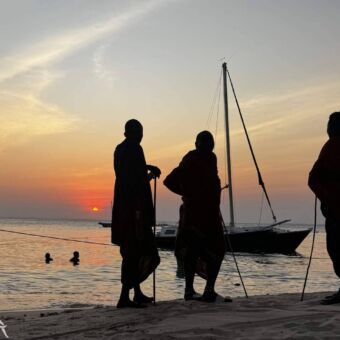 De silhouetten van drie mannen, waarschijnlijk Masaï, die op het strand staan tijdens zonsondergang, met een zeilboot in het water.