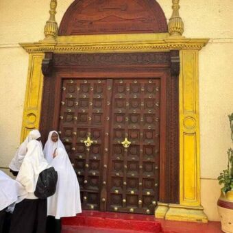 Houten deur met veel houtsnijwerk en koper accenten. Daarnaast staan drie vrouwen in witten gewaden