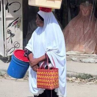 In lang gehad geklede vrouw met een doos op haar hoofd een tas in de en hand en emmers in de andere
