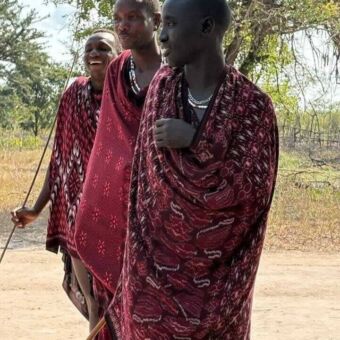 Drie masai mannen op een rij achter elkaar in kleurrijke gewaden. De achterste lacht hard