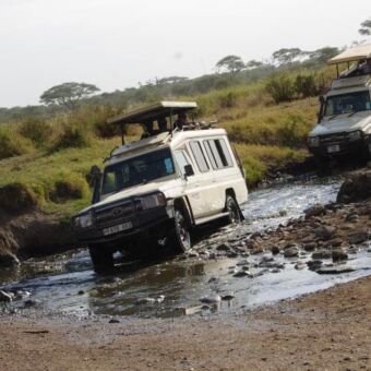 Zand weg en daarachter een riviertje waar een safariauto doorheen rijdt