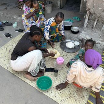 twee vrouwen en een paar kinderen zitten op een kleed voor hun huis te eten