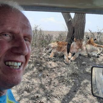 man voorin de safariauto en door het zijraam zie je vlakbij 3 leeuwen onder de boom liggen
