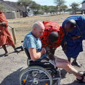 Drie Masai mannen waarvan twee voorovergebogen bij een man in een rolstoel