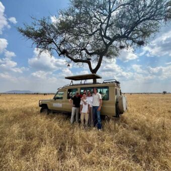 Familie poseert voor hun safariauto onder een grote acaciaboom op de vlaktes van de Serengeti