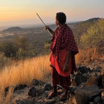 Een Maasai-man in traditionele rode kleding kijkt uit over de savanne tijdens zonsondergang